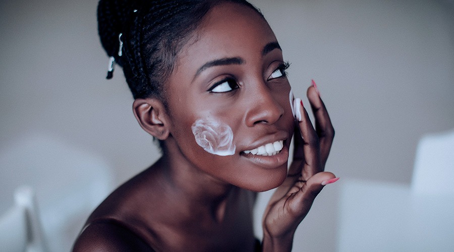 Plano fechado de uma mulher negra de perfil, sorrindo, passando creme branco no rosto