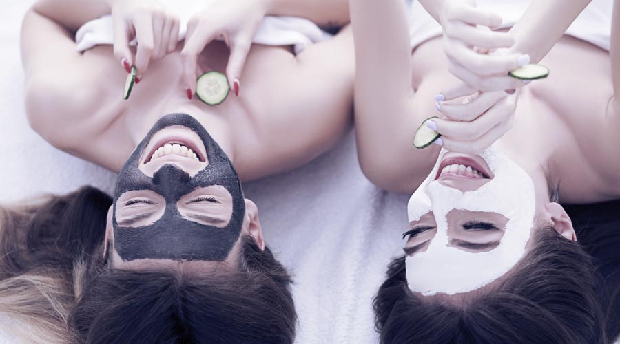duas amigas deitadas na cama, uma com uma máscara facial de argila preta e outra com argila branca.