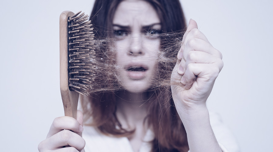 Uma mulher segura uma escova. Em primeiro plano o foco está nos vários cabelos acumulados na escova, enquanto ao fundo a mulher faz uma expressão de desespero.