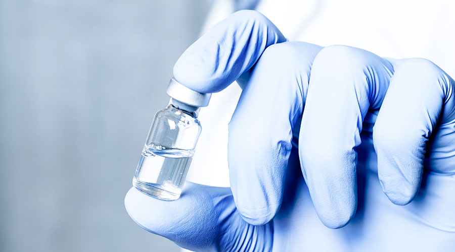Na imagem, mão com luva azul segura um frasco de vacina meio cheio.