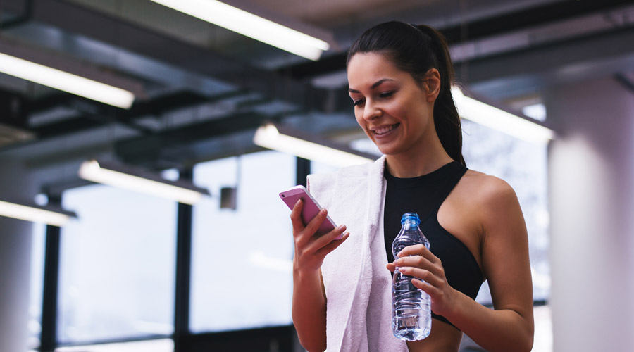 jovem mulher na academia, estabelecendo rotina saudável com um smartphone em uma mão e uma garrafa de água na outra