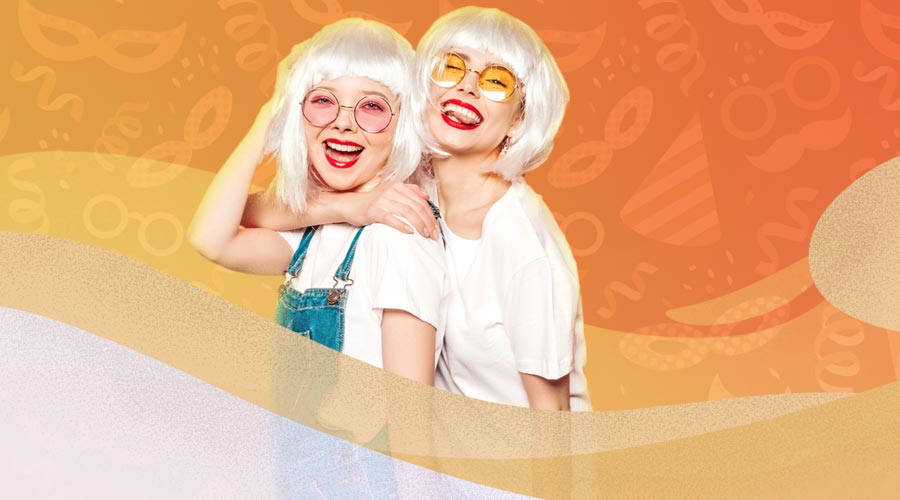 Uma das Dicas de Carnaval é usar roupas de tecidos como algodão. A imagem mostra uma composição em que duas meninas vestidas com roupas confortáveis usam peruca e se abraçam felizes.