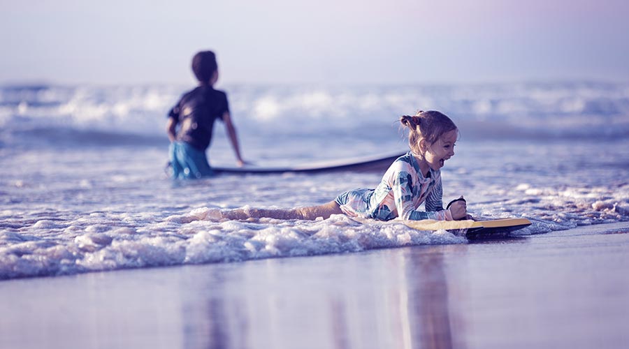 A imagem mostra uma criança dentro do mar, na parte rasa, praticando um esporte radical em cima de uma prancha.