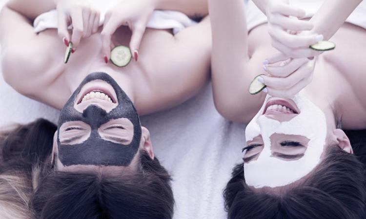 duas amigas deitadas na cama, uma com uma máscara facial de argila preta e outra com argila branca.